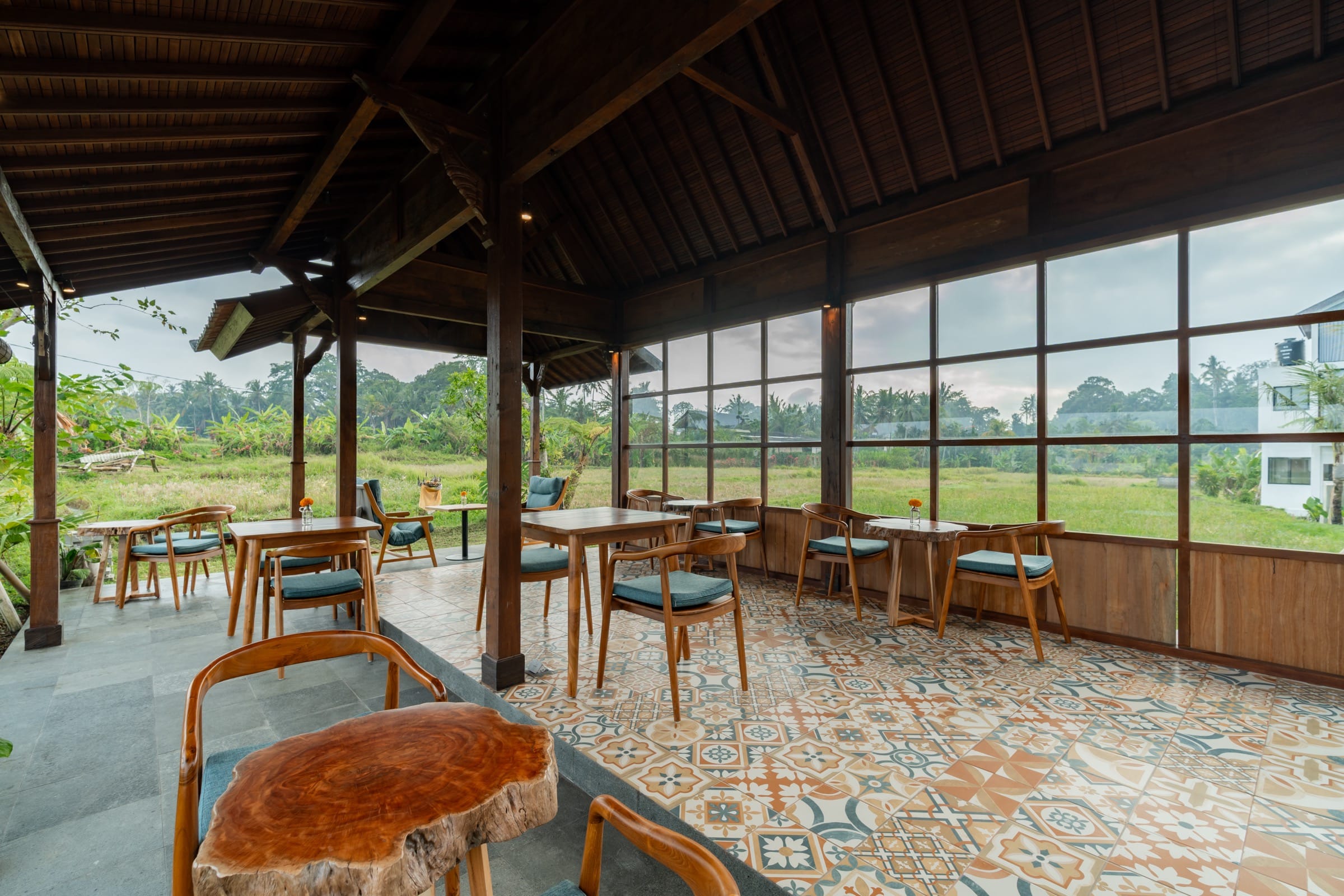 Tables and chairs on a patterned tile floor under the pavilion roof, with glass walls opening onto the rice field